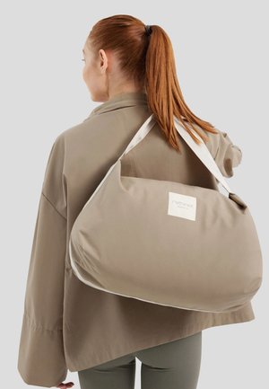 Bolso de tela beige con una forma suave y redondeada, que presenta un parche con el logo en blanco y correas largas y blancas en contraste, llevado sobre un abrigo gris.