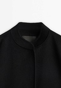 Manteau noir en tissu texturé, avec col montant et design minimaliste. L'avant ne présente aucun agencement ou ornement visible.