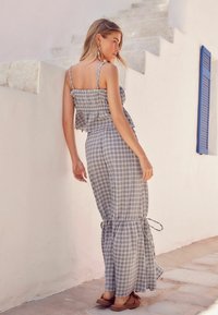 Abito maxi a quadri grigi con spalline volantee, vita arricciata e orlo con volant. Indossato con scarpe slip-on marroni su un pavimento in pietra.