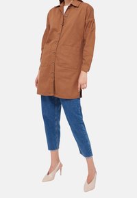 Hnědá košile s límcem, zapínáním na knoflíky, dvěma předními kapsami a ohrnutými rukávy, spárovaná se světle modrými džínami a béžovými lodičkami.