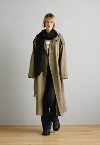 Trench coat bej cu guler supradimensionat și accesorii negre, asortat cu o eșarfă neagră, o bluză cu dungi, pantaloni negri și pantofi chunky negri.