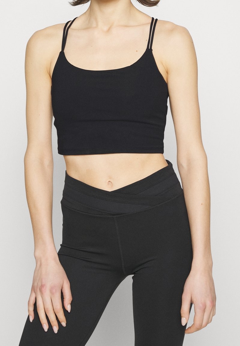 Femme portant un débardeur court noir à fines bretelles doubles et un legging noir taille haute avec une ceinture croisée.