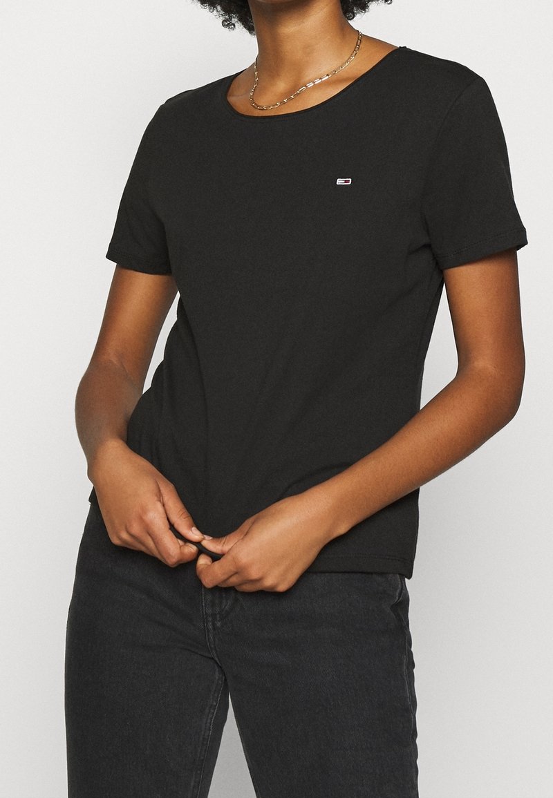 Žena ve fitted černém tričku s krátkým rukávem a malým obdélníkovým logem na hrudi, doplněném černými džínami.