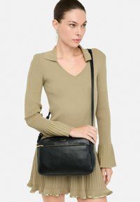 Sac en cuir noir porté à l'épaule avec une finition texturée, doté d'une poche zippée à l'avant et d'une bandoulière réglable, portée par un modèle en robe vert clair.