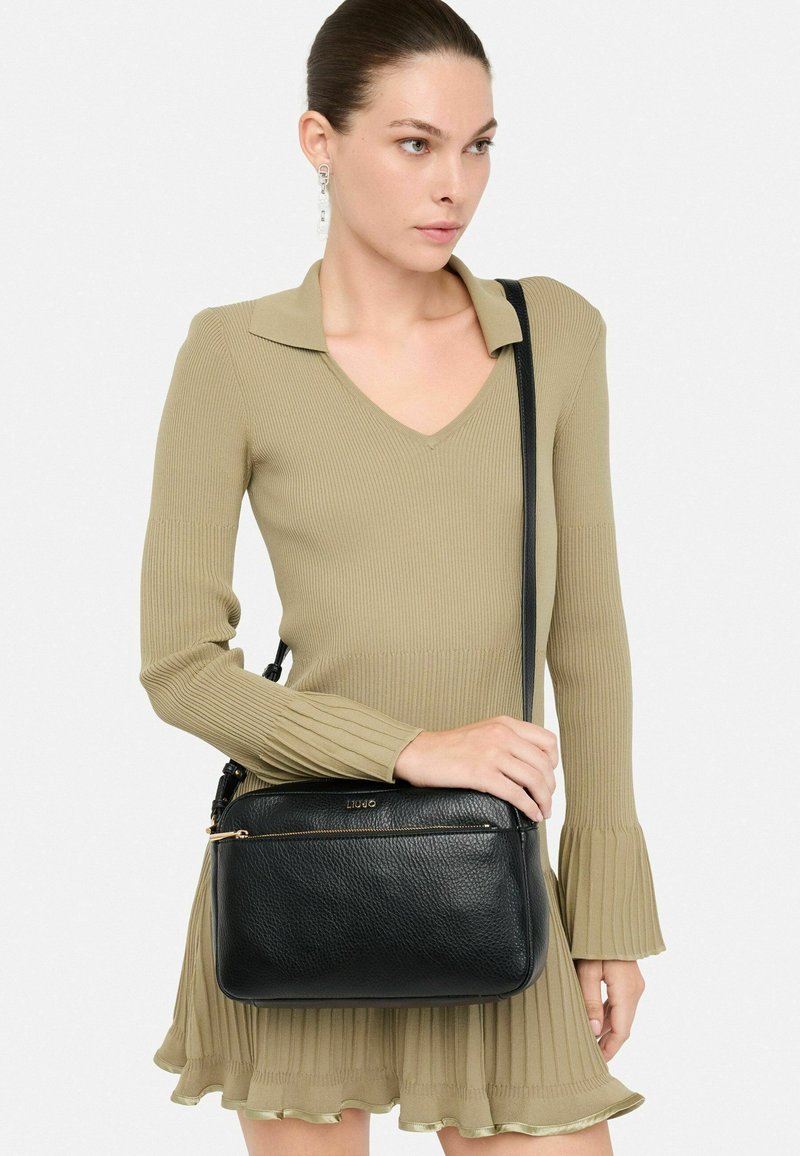 Sac en cuir noir porté à l'épaule avec une finition texturée, doté d'une poche zippée à l'avant et d'une bandoulière réglable, portée par un modèle en robe vert clair.