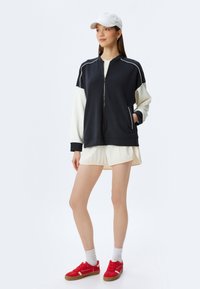Marinblå zip-jacka med vita ärmar, försedda med sidofickor. Matchas med krämfärgade shorts och röda sneakers. Vit keps fulländar looken.