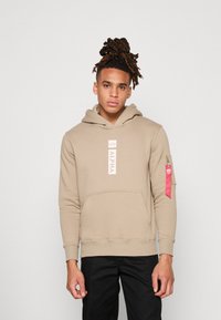 Beige hettegenser med frontlomme, snørehette og en vertikal hvit logo-lapp. Høyre erme har en rød tekstilaccent med glidelås.