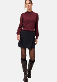 Femme portant un haut bordeaux à manches longues avec des manches plissées, une jupe portefeuille noire, des collants transparents et des bottes marron foncé jusqu'aux genoux sur fond blanc.