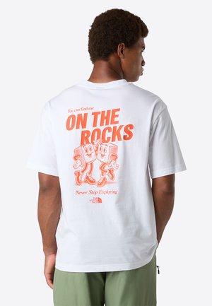 Mand iført hvid t-shirt med orange grafik af to tegneserieisterninger og teksten "ON THE ROCKS" på ryggen, parret med grønne shorts.