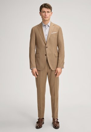 Mężczyzna w beżowym garniturze, jasnoszarej koszuli i brązowych eleganckich butach, stoi na jasnym jednolitym tle, zwrócony przodem.