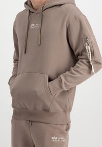 Sweat à capuche en coton marron avec une poche à l'avant, des poignets côtelés et des cordons de serrage réglables. Comprend un logo et une poche zippée sur la manche.