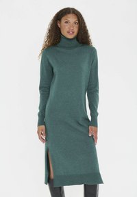 Femeie îmbrăcată cu o rochie tricotată verde, cu mâneci lungi și guler înalt, având o fereastră laterală, stând în fața unui fundal alb simplu.