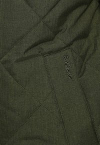 Tecido verde em espinha de peixe acolchoado com superfície texturada. A marca "Barbour" está discretamente bordada ao longo de uma das bordas.