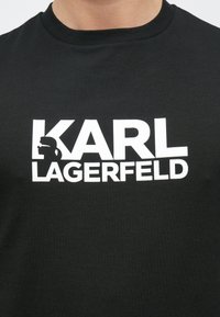 Fekete pamut póló, amelyen vastag fehér felirat olvasható: "KARL LAGERFELD", grafikusan kiemelt sziluettel. Klasszikus környakas kivitel.