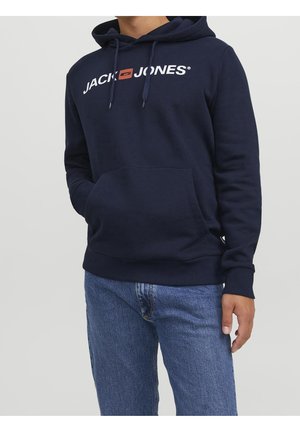 Mężczyzna ubrany w granatową bluzę z kapturem z logo „Jack & Jones” i niebieskie dżinsy, stojący z jedną ręką w kieszeni bluzy.