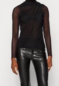 Svart långärmad topp med ett högre halsringning, i en genomskinlig material, med volanger fram och en figurnära design. Glansiga svarta byxor kompletterar den.