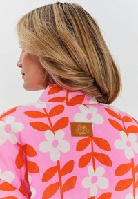 Ružová bunda s veľkými bielymi kvetmi a oranžovými listami. Obsahuje hnedú koženú štítku s textom "Butter Toast Mornings".