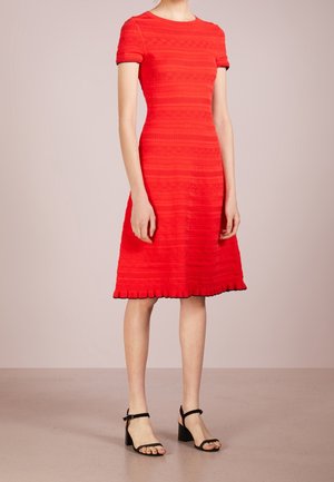Femme portant une robe rouge texturée, courte aux manches, longueur genoux, associée à des sandales noires à talons blocs avec bride à la cheville, debout sur un sol uni.