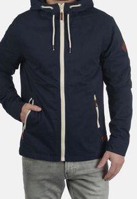 Veste bleu marine avec capuche, fermeture éclair blanche et cordons. Comprend des accents en cuir beige et des poches latérales. Texture lisse, design léger.