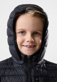 Giacca invernale nera con cappuccio. Presenta una texture trapuntata e una chiusura con zipper. La persona ha i capelli chiari e un sorriso smagliante.