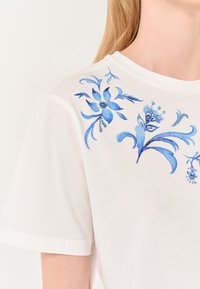 T-shirt bianco in cotone con motivi floreali blu, comprese decorazioni intricate e dettagli, vicino all'area del collo e delle spalle.