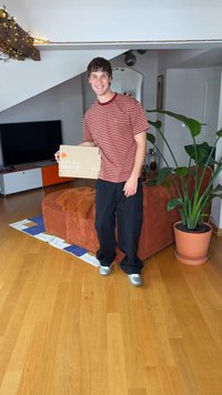 Papæske mærket "Elsk mig, bær mig" holdt af en mand i en rød- og hvidstribet skjorte og sorte bukser, stående i et lyst rum med planter.