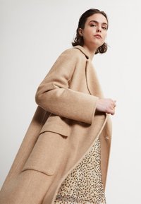 Abrigo de mezcla de lana beige con textura de espiga, solapas con muesca y bolsillos laterales, sobre un vestido de patrón salpicado de oro.