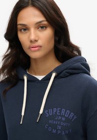 Sudadera con capucha de color azul marino con un logotipo frontal en púrpura, cordones color crema y una tela suave y texturizada. Presenta un bolsillo tipo canguro.