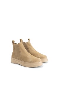Een paar beige suède Chelsea-boots met elastische zijpanelen en dikke rubberen zolen op een witte achtergrond.