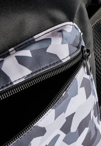 Sac à dos en tissu texturé noir avec un motif camouflage gris et blanc. Comprend une poche zippée avec des coutures contrastées.