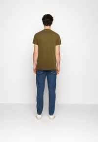 Tricou scurt cu mânecă scurtă de culoare verde măsliniu, cu guler rotund, asortat cu blugi albaștri, văzut din spate pe un fundal alb simplu.