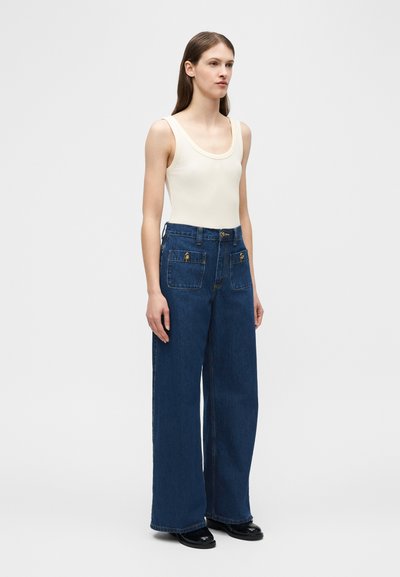 Νεαρή γυναίκα που φοράει μπεζ τοπ και παντελόνι ψηλόμεσο με φαρδιά μπατζάκια σε σκούρο μπλε χρώμα, με κουμπιά μπροστά στις τσέπες, στέκεται μπροστά σε λευκό φόντο.