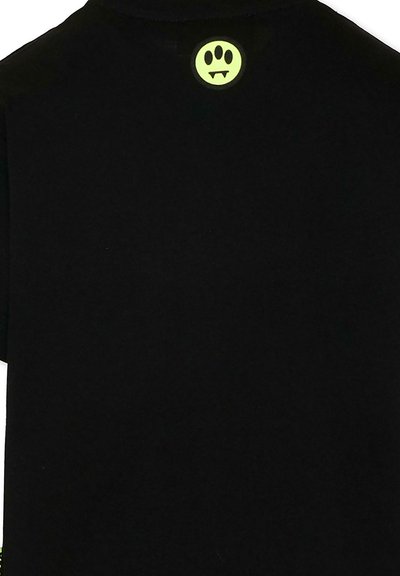 Czarna koszulka z małą, neonowożółtą buźką z wampirzymi kłami umieszczoną w pobliżu górnego kołnierza na plecach.