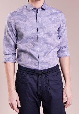 Mann trägt ein hellviolettes, camouflagegemustertes Hemd mit Knöpfen, die Ärmel sind aufgekrempelt, dazu dunkelblaue Jogginghosen mit Kordelzug.