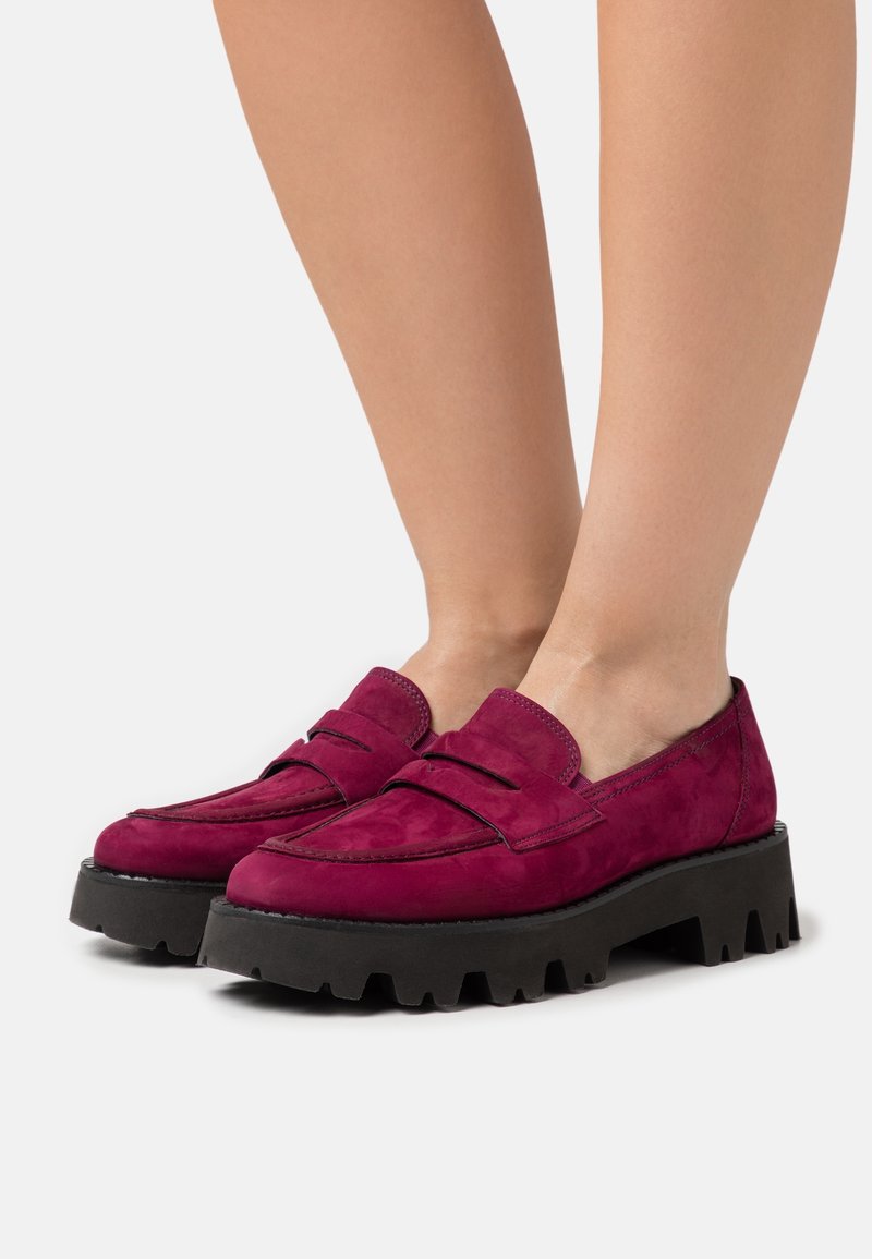 Paire de mocassins en daim magenta avec des semelles épaisses noires à crampons portés pieds nus sur un fond blanc uni.