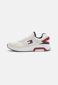 Zapatilla blanca con parte superior de ante y tejido, acentos en azul marino, lengüeta trasera roja y un logo de marca en el lateral. Presenta una suela acolchada para mayor comodidad.