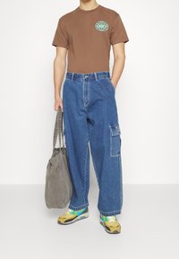 Hnědé tričko s logem "OBEY", modré denimové cargo kalhoty s bočními kapsami, šedá taška přes rameno a žluté tenisky s akcenty v modrozelené barvě.