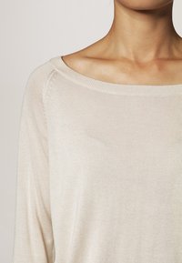 Světle béžový pletený svetr s lodním výstřihem. Má dlouhé rukávy a hladkou texturu, která zdůrazňuje jednoduchý a uvolněný design.