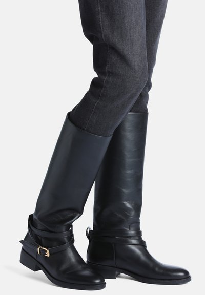 Personne portant des bottes en cuir noires montantes avec des sangles enveloppantes et une boucle dorée, associées à un jean en denim foncé.