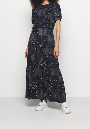 Woman wearing a black patchwork-patterned maxi dress with short sleeves and white sneakers, standing against a plain background.