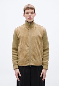 Taninavärvi jakk, millel on seemisnahast esikülg ja soonikkoes varrukad, kõrge krae luku ja küljetaskutega. Lihtne, kaasaegne disain.