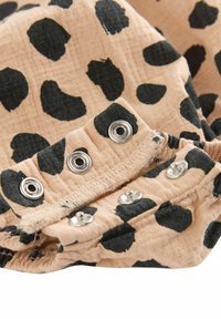 Ljusbeige tyg med svarta leopardfläckar, som har silverfärgade tryckknappar längs kanten, vilket skapar ett strukturerat, lekfullt mönster.