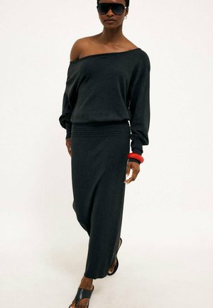 Woman wearing black off-shoulder top and long skirt, black sunglasses, red and black bracelets, walking barefoot on a white background.