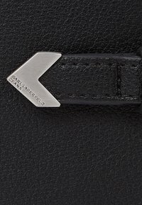 Juodas tekstūruotas oda su sidabriniu metaliniu akcentu, formuojančiu chevroną. Metaliniai elementai turi įspaustą užrašą "Karl Lagerfeld Jeans".