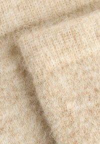 Beige gestrickter Stoff mit einer weichen, flauschigen Textur. Das Muster zeigt eine lockere Webart, die ein warmes, gemütliches Erscheinungsbild erzeugt.