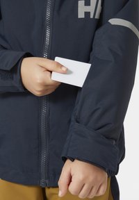 Námořnická nepromokavá bunda s předním zipem a nastavitelnými manžetami; ruka drží bílou kartu v kapse na rukávu, což ukazuje na funkčnost.