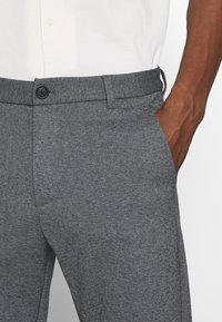 Pantalon gris texturé avec une coupe droite, comportant une fermeture à bouton, des poches inclinées et une taille avec un détail à cordon.