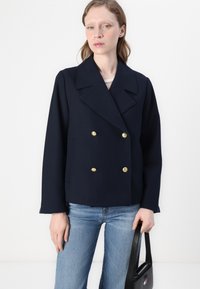 Mørkeblå dobbeltradet jakke med store guldknapper, brede revers og cropped design, parret med lyseblå jeans og en sort håndtaske.