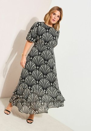 Vrouw draagt een enkellange jurk met zwart-wit patroon en pofmouwen, en zwarte open teenschoenen, staand tegen een effen muur.