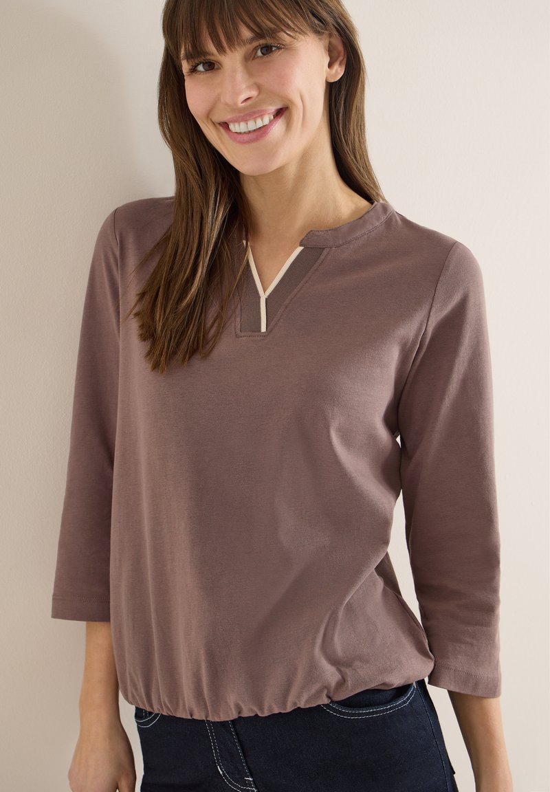 Femme souriante aux longs cheveux bruns portant un haut mauve à manches longues avec un col en V et un ourlet élastique, debout contre un mur clair.